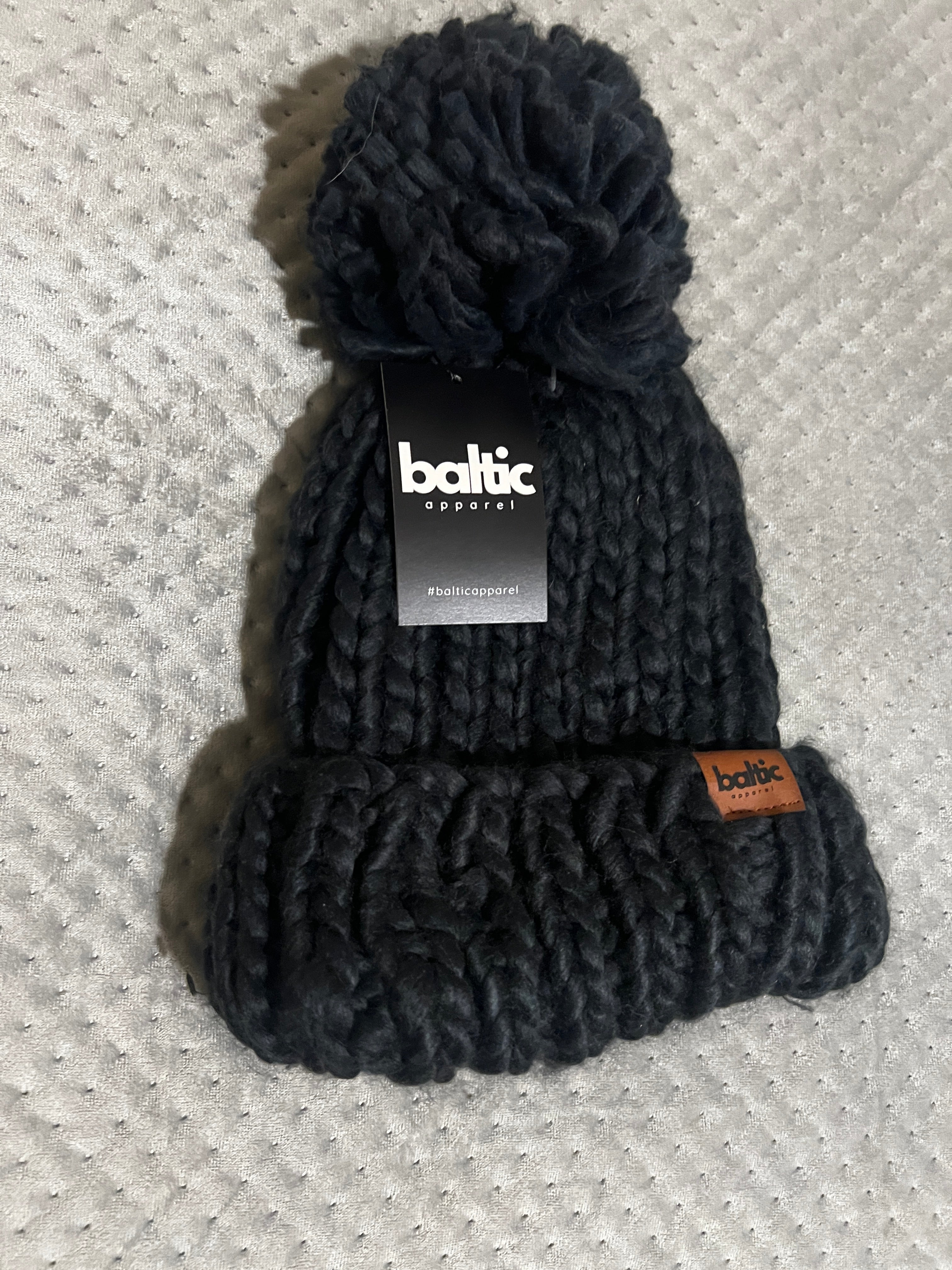 Black knitted headband with pom-pom on a textured gray surface, featuring a Baltic brand label.