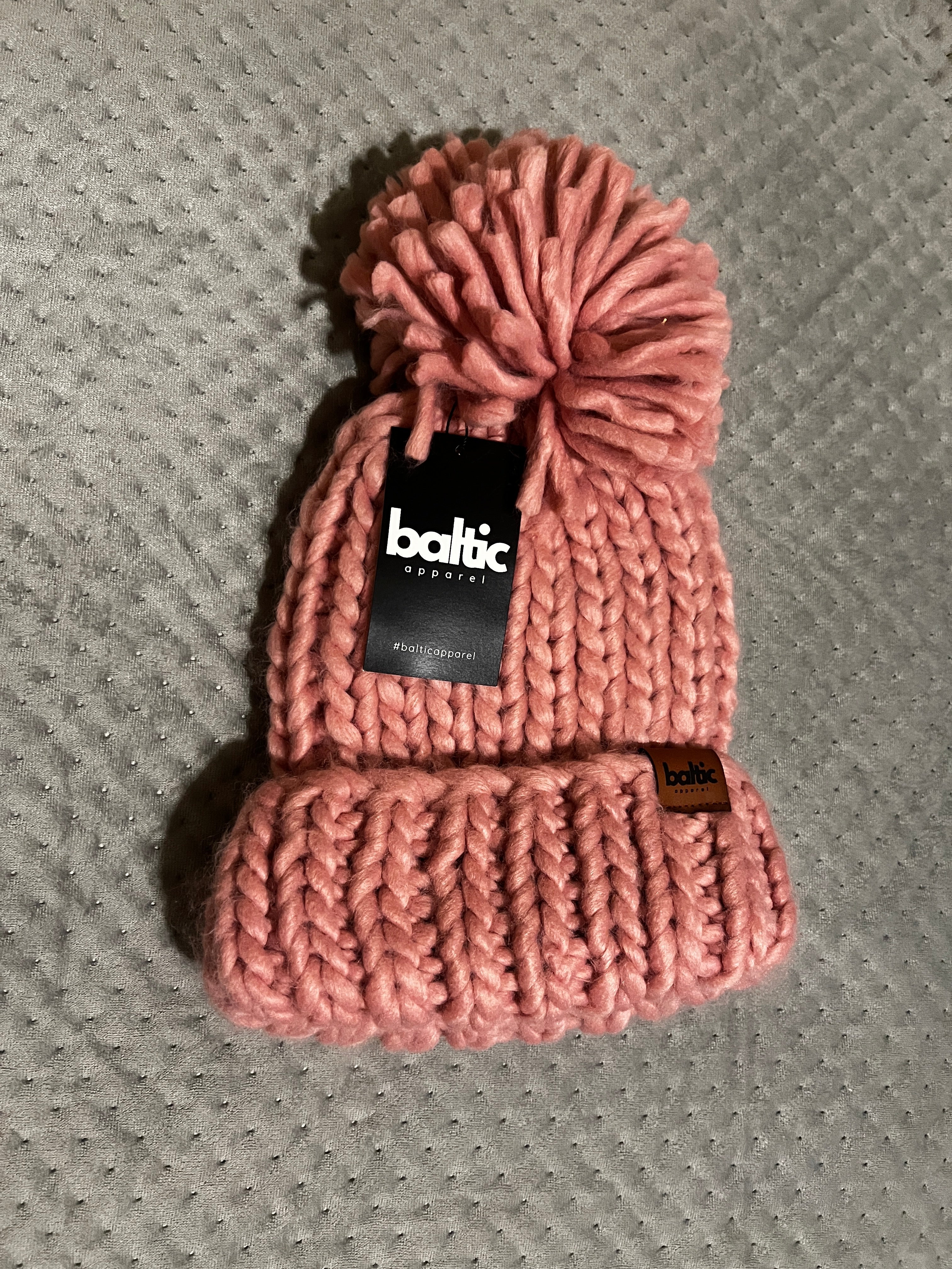 Pink knitted headband with a pom-pom on a textured gray surface, featuring a brand label.