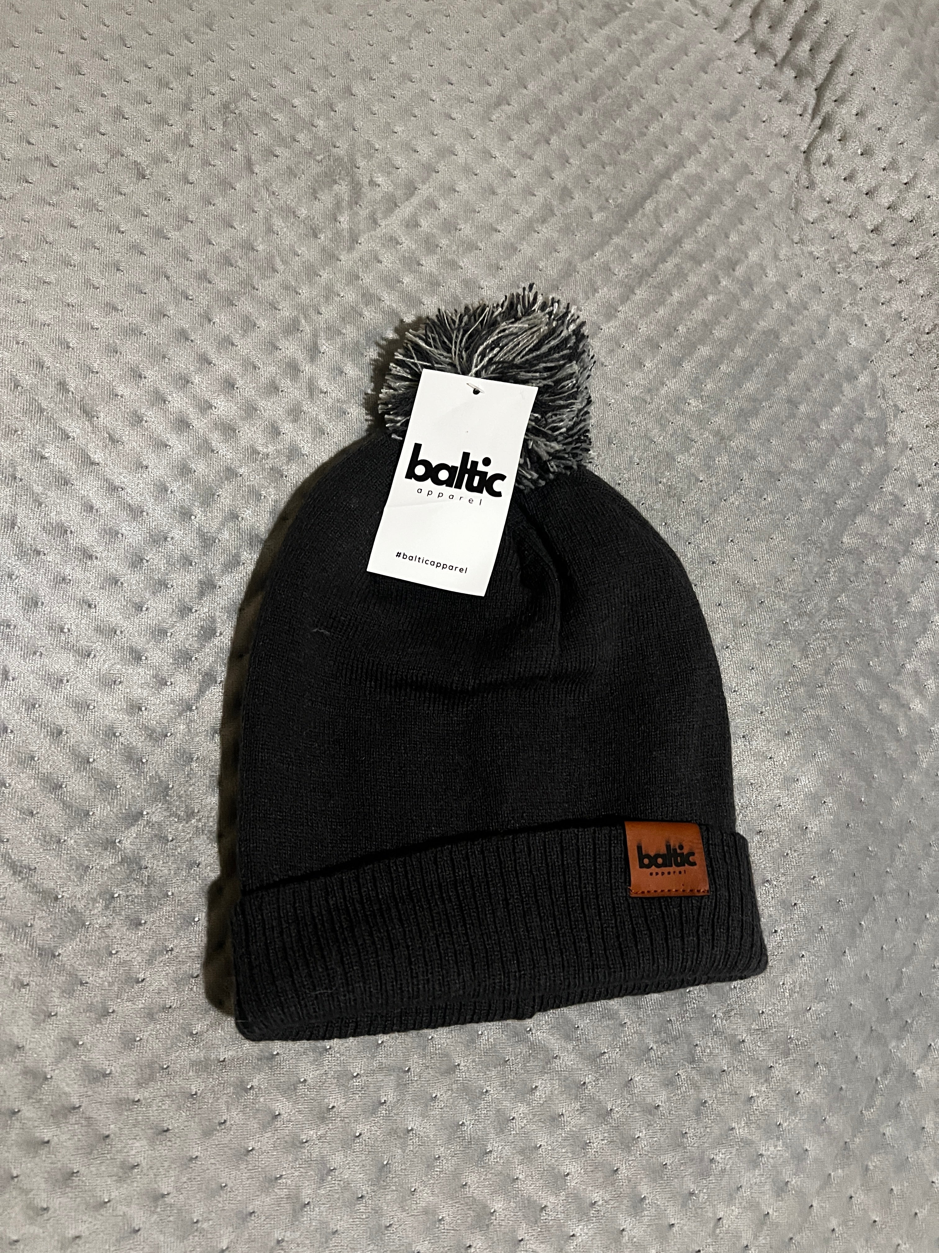 Black beanie with pom-pom and brand tag on a textured gray surface