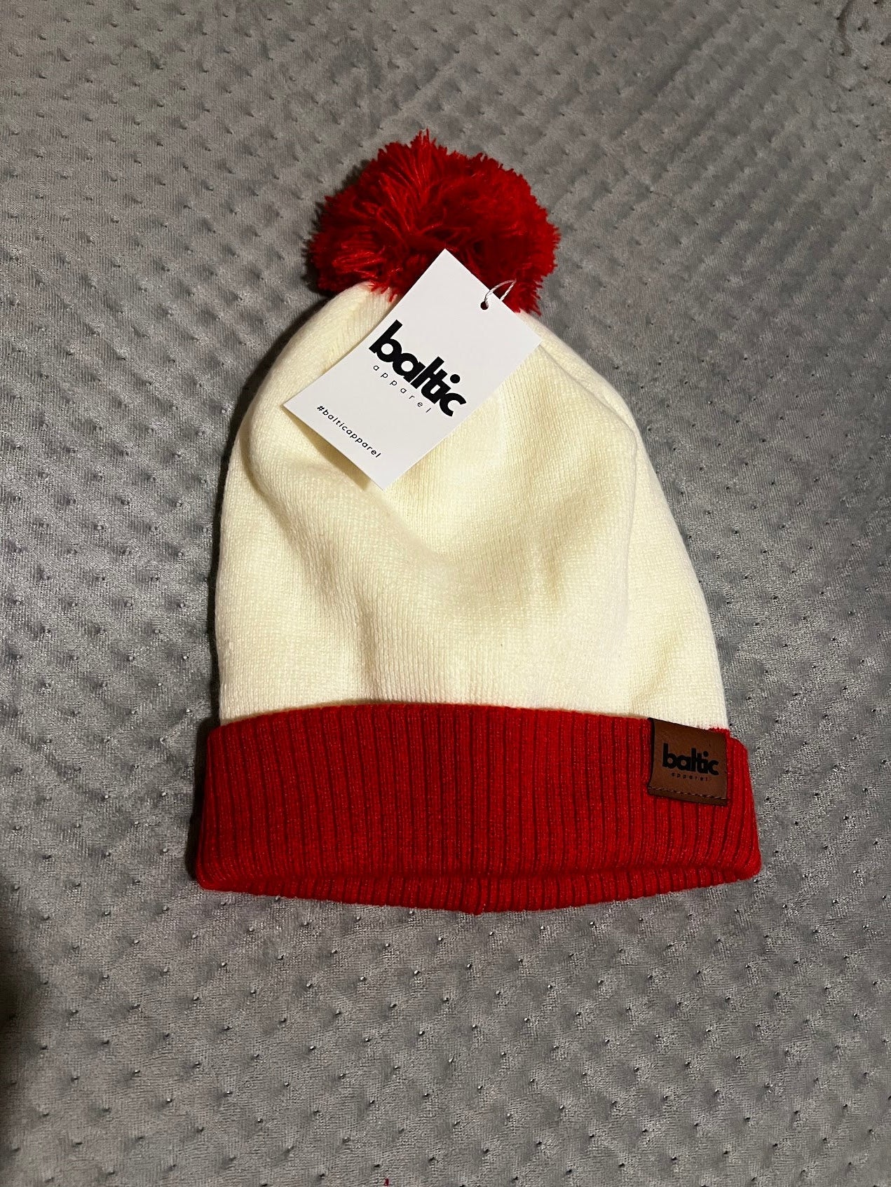 White beanie with red pom-pom and band on a textured gray surface, featuring a Baltic brand tag.