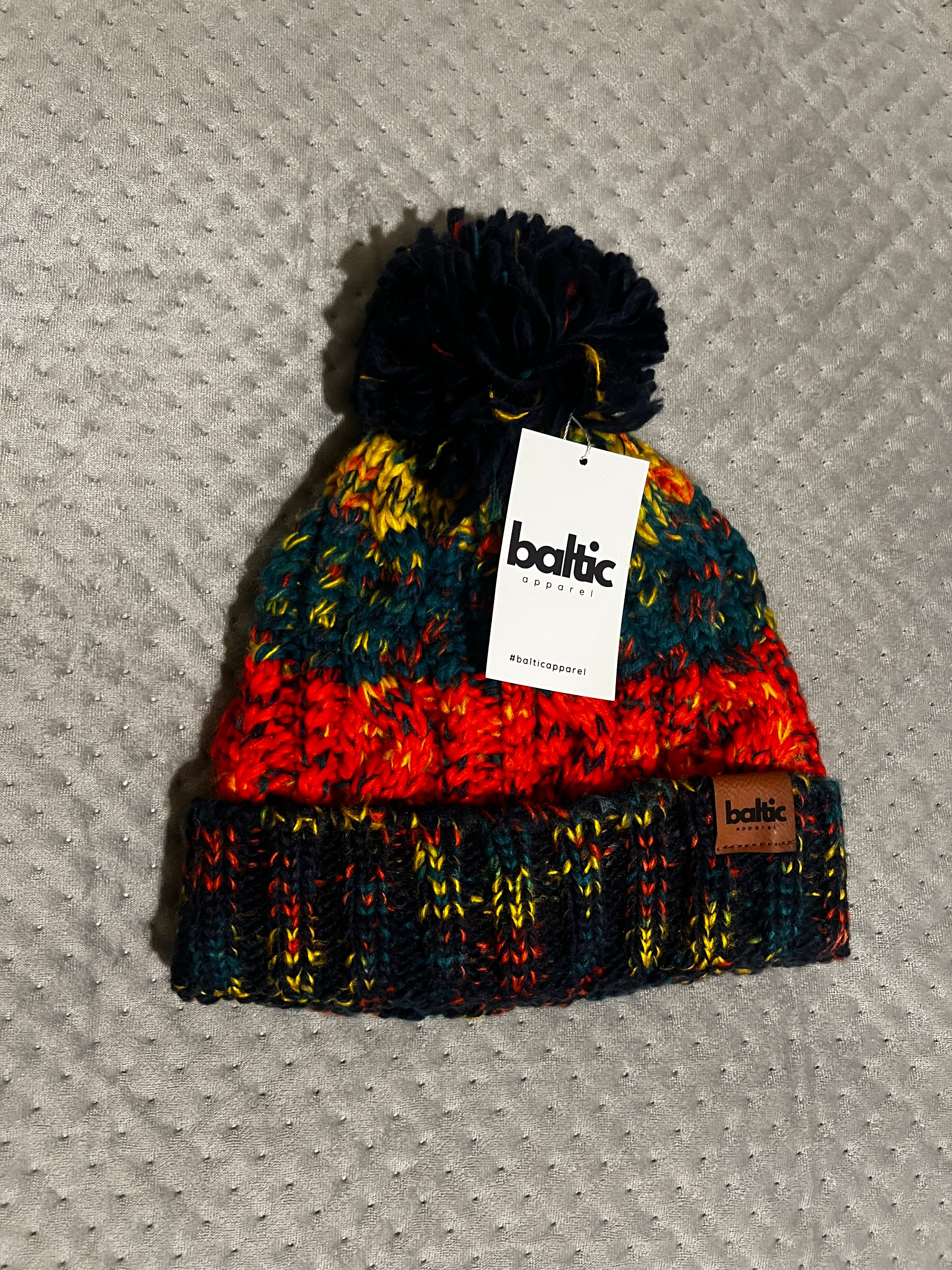 Colorful knit beanie with a pom-pom and Baltic label on a textured gray surface